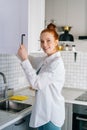 Side view of cheerful redhead young woman opening door of kitchen cabinet at light kitchen room. Royalty Free Stock Photo