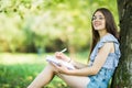 Side view of concentrated brunette woman in eyeglasses sitting near the tree in park and writing something on notebook