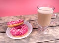 Side view of a cup of cofee with milk and two pink donuts on a white plate standing on wooden planks on pink background Royalty Free Stock Photo