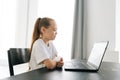 Side view of cute blonde little girl with broken hand wrapped in white plaster bandage sitting at table with laptop Royalty Free Stock Photo