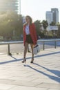 Side view of an elegant woman wearing sunglasses, red jacket, skirt and holding a white handbag while walking in the street in a Royalty Free Stock Photo