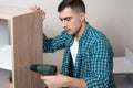 Side view of European man in shirt with electric screwdriver in hand engaged in the Assembly of furniture