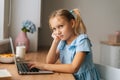 Side view of exhausted elementary child girl using laptop sitting at home table with snack by window, looking at camera. Royalty Free Stock Photo