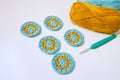 Side view of five round crochet motifs in turosque and yellow colors, a hook and skeins of yarn on smooth white surface