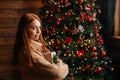 Side view of flirty redhead young woman holding Christmas gift box on background of xmas tree. Royalty Free Stock Photo