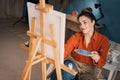 side view focused ethnic woman in casual wear sits on chair draws on canvas with colorful paints and brush in small room