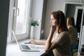 Side view of focused female operator talking using headset and consulting client sitting at window sill with laptop from