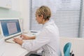 Side view of focused middle-aged woman general practitioner in white coat using laptop computer writing notes at Royalty Free Stock Photo