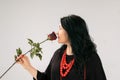 Side View of Full Figured Woman in Black Dress and Ethnic Necklace Sniffing Red Rose on Grey Background in Studio Royalty Free Stock Photo