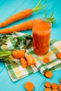 side view of glass of carrot juice with cut and sliced carrots on cloth decorated with leaves on blue background