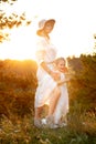 Side view of gorgeous family wearing white dresses, standing near pines in forest lit up by sunset in summer. Vertical.