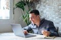 Side view handsome young businessman in eyewear working with computer remotely, sitting at wooden table in office. Pleasant happy