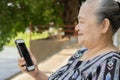 Side view of happy Asian elder senior grey haired woman sitting outdoor and holding mobile phone Royalty Free Stock Photo