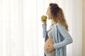 Side view of happy young pregnant lady standing by the window and eating green apple Royalty Free Stock Photo