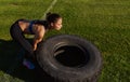 Side view image of athlete strong fitness woman doing tire-flip outdoors on green grass in stadium. Copy space for advertising.