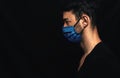 Side view image of Caucasianman wearing medical blue mask on the face during virus pandemic lockdown posing on the black wall with Royalty Free Stock Photo