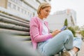 Side view image of pretty blonde young woman wearing blue jeans, white t-shirt and pink jacket, smiling and sending messages on Royalty Free Stock Photo