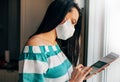 Side view image of young woman in white medical mask next the window and working on the tablet. Female with protective face mask