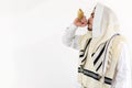 Side view of a Jewish man in tallith blowing the Shofar against a white background.