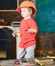 Side view kid standing in workshop. Blond boy in orange helmet holding spanner and handsaw. Little repairman posing for Royalty Free Stock Photo