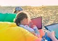 Side view of little girl sitting along other kids in colorful bag chair and drawing landscape with pastel at the beach