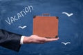 Side view of man`s hand facing up and holding brown travel case against blackboard with chalk birds and title `Vacation Royalty Free Stock Photo