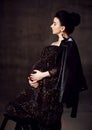 Side view of noble pregnant noble woman in black off-shoulder dress and leather jacket stands holding hand at her belly Royalty Free Stock Photo