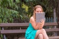 Side view of pleased blonde girl in blue dress sitting on bench with reading book in park. Fascinating reading literature outdoors Royalty Free Stock Photo