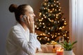 Side view portrait of adorable beautiful woman with bun hairstyle wearing white shirt sitting on table near christmas tree, Royalty Free Stock Photo