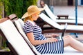 Side view portrait of beautiful happy young adult freelancer woman in hat and dress is sitting on cozy daybed with laptop on Royalty Free Stock Photo