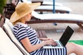 Side view portrait of beautiful serious young adult writer woman in hat and dress is sitting on cozy daybed with laptop on Royalty Free Stock Photo