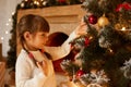 Side view portrait of charming little girl with pigtails decorating Christmas tree alone, wearing white sweater, standing in Royalty Free Stock Photo