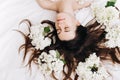 Side view portrait of dreamful young brunette woman with closed eyes and sincere smile lying in comfy white bed, spring flowers