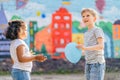 Side view portrait of happy little blond boy giving blue balloon for cute little hispanic curly preschool girlfriend, outdoor. Royalty Free Stock Photo