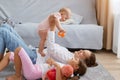 Side view portrait of happy woman wearing white shirt and jeans lying on floor near sofa with her daughters, raising baby, Royalty Free Stock Photo