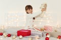 Side view portrait of little girl wearing white sweater holding soft toy dog, sitting on bed with Christmas decoration and garland Royalty Free Stock Photo