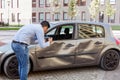 Side view portrait of man wearing jeans and shirt making photo of damaged car after auto accident, photographing dents and