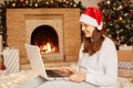Side view portrait of smiling woman wearing white sweater and santa claus hat working with her laptop computer, posing in festive Royalty Free Stock Photo