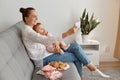 Side view portrait of smiling woman with bun hairstyle wearing white shirt and jeans sitting on sofa with her baby daughter and Royalty Free Stock Photo