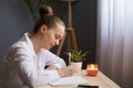 Side view portrait of young beautiful focused woman writing down notes while sitting at table in office in front of the window, Royalty Free Stock Photo