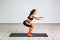 Side view portrait of young sporty healthy beautiful woman in black top and orange leggings doing squatting with elastic Royalty Free Stock Photo