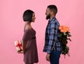 Side view of positive black couple with flowers and gift for Valentine& x27;s Day looking at each other over pink background