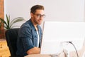 Side view of positive young designer male in stylish glasses working on desktop computer sitting at desk at home office Royalty Free Stock Photo