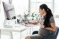 View of pregnant woman sitting behind table with computer, keyboard and document tray, writing notes and holding belly Royalty Free Stock Photo