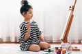 Side view of a pretty little girl painting with a paintbrush on the paper on an easel at home. A cute child sitting on the floor Royalty Free Stock Photo