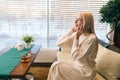 Side view of pretty young woman smiling talking on mobile phone sitting on sofa at table with tea by window. Happy Royalty Free Stock Photo