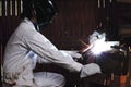 Side view of professional welder in white uniform with protective helmet welding steel with spark in workshop. Industrial worker c