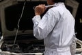 Side view of professional young mechanic man in uniform holding wrench against car in open hood at the repair garage. Royalty Free Stock Photo