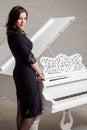 Side view profile of sensual brunette woman in lace black classic dress standing near white piano and looking at camera