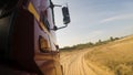 Side view of red truck cab moving on dusty unpaved rural road along green trees ahead. Scene. Huge lorry riding on